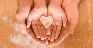 Charitable giving webinar open graph is an image of a child's hands, being held underneath by a parent. In the childs hands they are holding a piece of dough shaped into a heart. Their hands are covered in flour and there is flour on the counter in the background of the photo.